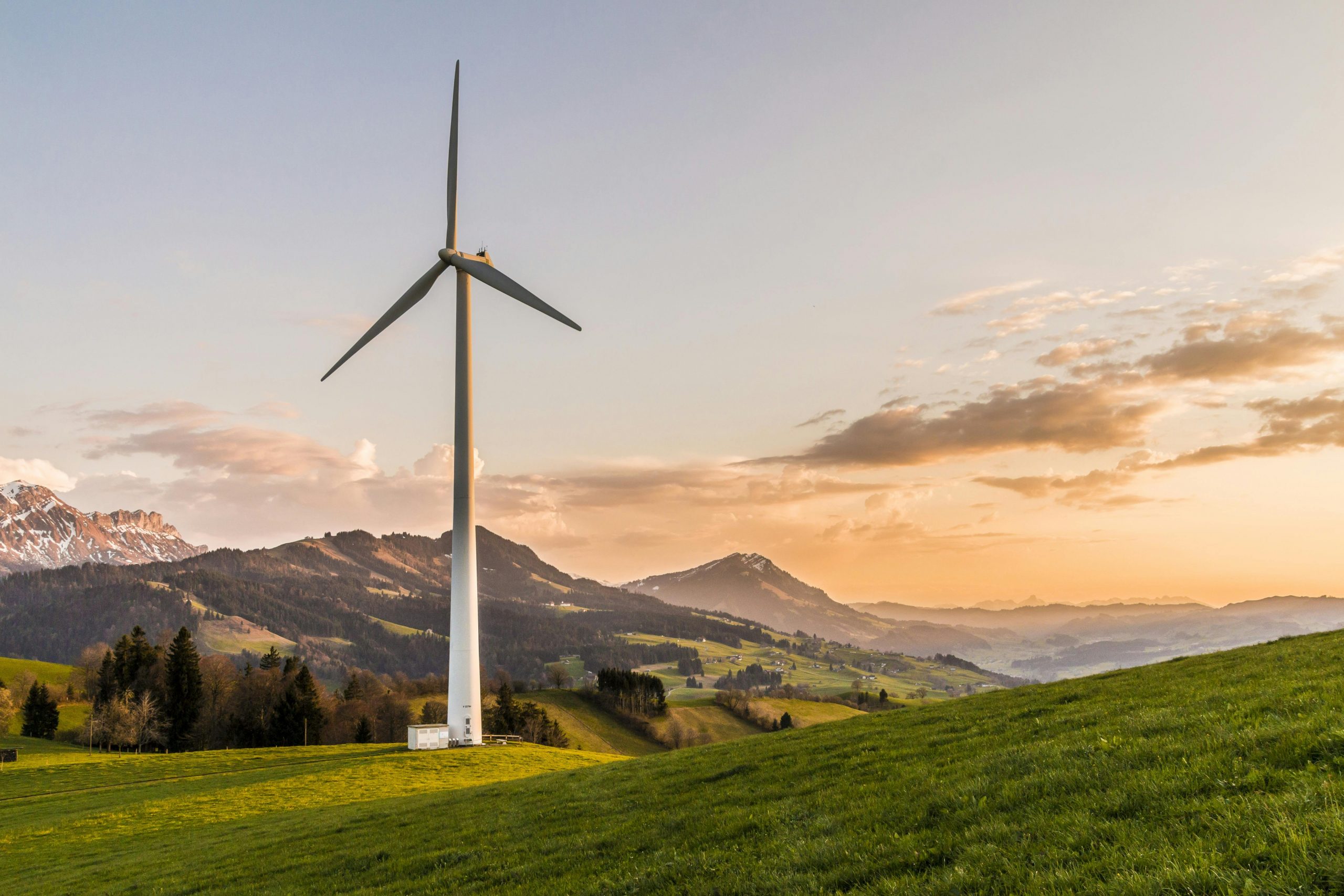 Das Bild steht in Zusammenhang mit Mediation und Konfliktlösung durch die Kanzlei Ponschab & Partner und zeigt folgendes: Eine große Windkraftanlage steht auf einem grünen Hügel mit Bäumen und Bergen im Hintergrund unter einem teilweise bewölkten Himmel bei Sonnenuntergang. Bei Fragen zu Konflikten und Konfliktlösung wenden Sie sich bitte an uns unter info@ponschab-partner.com