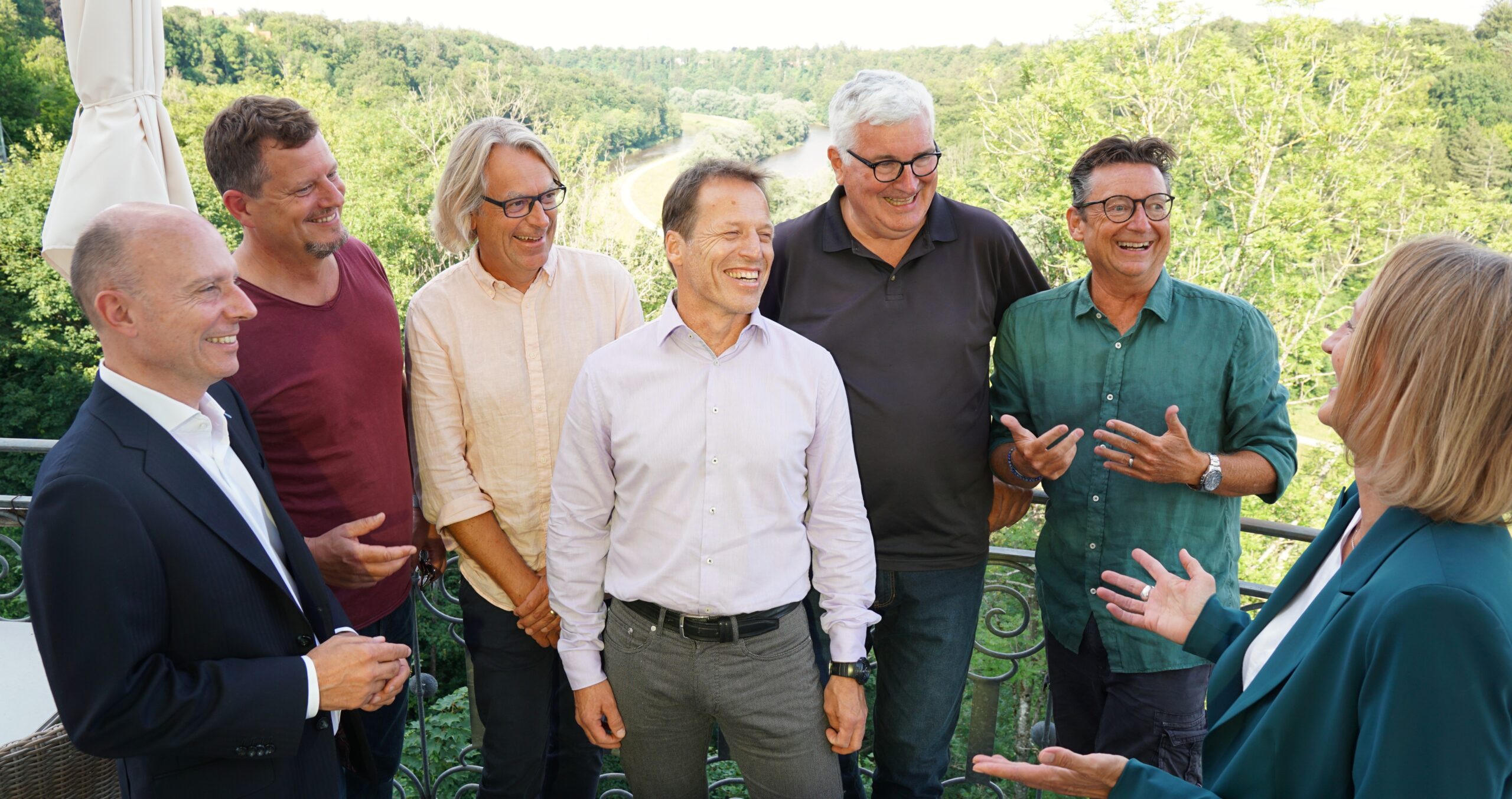 Das Bild steht in Zusammenhang mit Mediation und Konfliktlösung durch die Kanzlei Ponschab & Partner und zeigt folgendes: Eine Gruppe von sieben Personen, eine Frau und sechs Männer, steht als Teil des Mediations-Teams draußen auf einer Terrasse. Sie unterhalten sich angeregt, lächeln und lachen. Im Hintergrund ist eine üppige, grüne Landschaft unter einem klaren Himmel zu sehen. Bei Fragen zu Konflikten und Konfliktlösung wenden Sie sich bitte an uns unter info@ponschab-partner.com