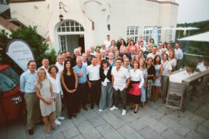 Das Bild steht in Zusammenhang mit Mediation und Konfliktlösung durch die Kanzlei Ponschab & Partner und zeigt folgendes: Eine große Gruppe von Menschen, sowohl Männer als auch Frauen, posieren gemeinsam im Freien auf einer Terrasse vor einem weißen Gebäude. Einige Personen stehen, während andere leicht geduckt davor stehen. In der Nähe sind Tische und Transparente zu sehen. Bei Fragen zu Konflikten und Konfliktlösung wenden Sie sich bitte an uns unter info@ponschab-partner.com