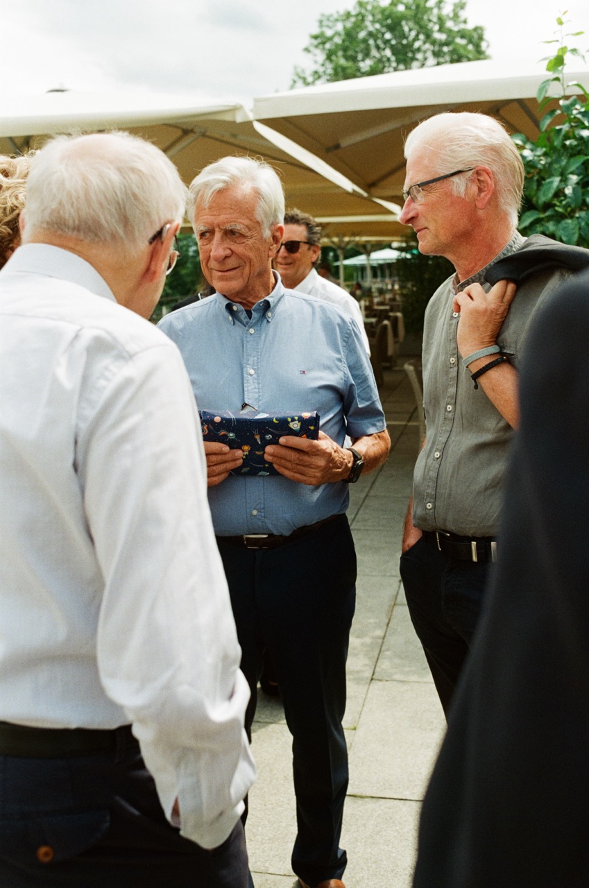 Das Bild steht in Zusammenhang mit Mediation und Konfliktlösung durch die Kanzlei Ponschab & Partner und zeigt folgendes: Eine Gruppe älterer Männer steht im Freien und unterhält sich. Ein Mann in der Mitte hält eine gemusterte Tasche in der Hand, ein anderer hat eine Jacke über der Schulter. Sie sind leger gekleidet und stehen auf einer gepflasterten Fläche mit Regenschirmen im Hintergrund. Bei Fragen zu Konflikten und Konfliktlösung wenden Sie sich bitte an uns unter info@ponschab-partner.com
