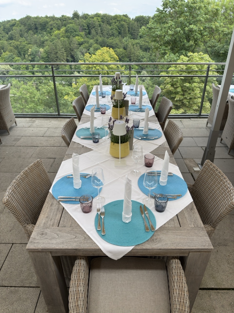 Das Bild steht in Zusammenhang mit Mediation und Konfliktlösung durch die Kanzlei Ponschab & Partner und zeigt folgendes: Ein langer Holztisch im Freien ist mit blauen Tischsets, weißen Servietten, Gläsern und Besteck für eine Mahlzeit gedeckt. Der Tisch blickt auf eine grüne, bewaldete Landschaft hinter einem Metallgeländer. Bei Fragen zu Konflikten und Konfliktlösung wenden Sie sich bitte an uns unter info@ponschab-partner.com