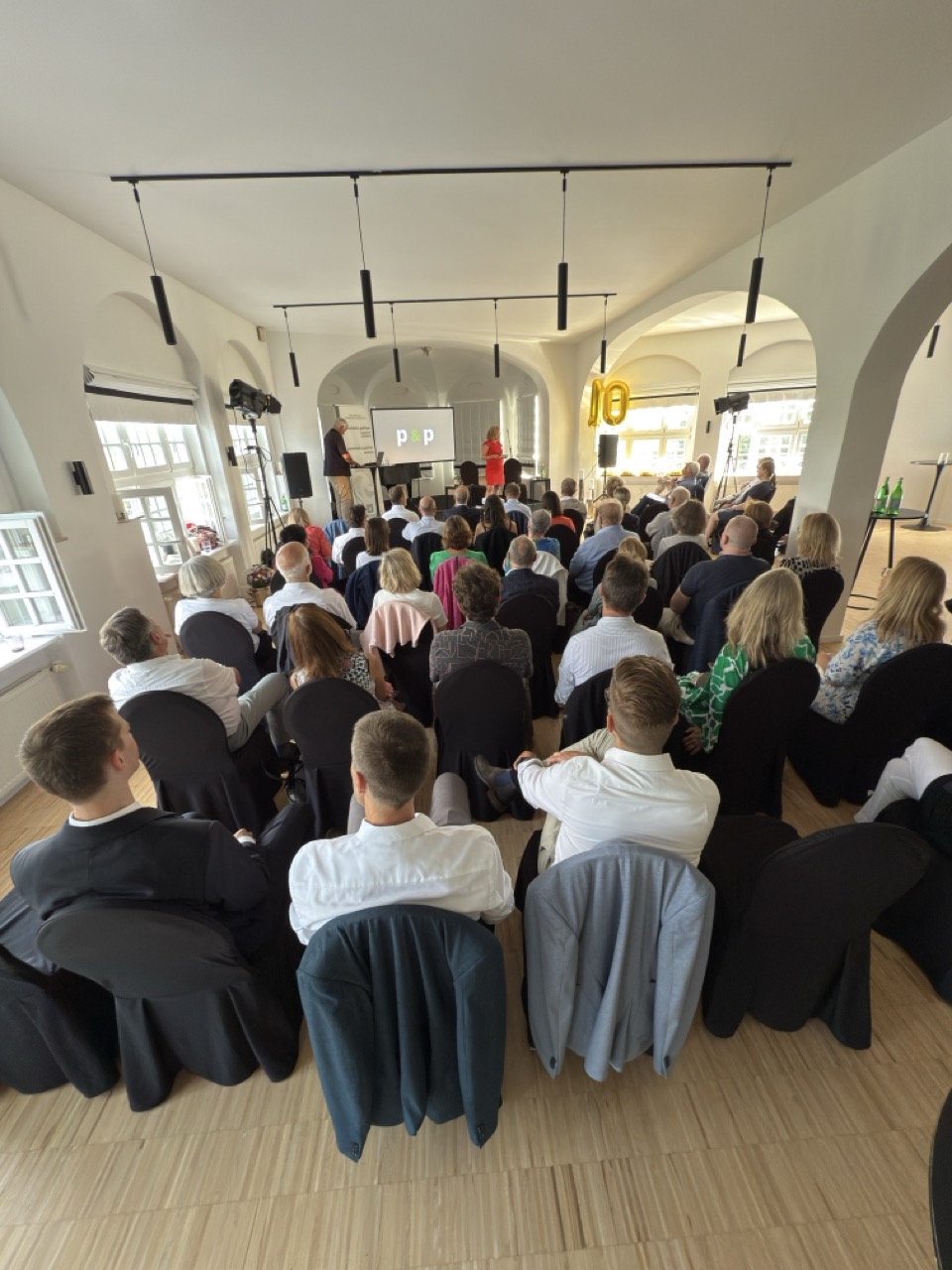 Das Bild steht in Zusammenhang mit Mediation und Konfliktlösung durch die Kanzlei Ponschab & Partner und zeigt folgendes: In einem hellen, modernen Raum mit großen Fenstern sitzt eine Gruppe von Menschen auf schwarzen Stuhlreihen vor einer Bühne, auf der Redner vor einem Präsentationsbildschirm stehen. Bei Fragen zu Konflikten und Konfliktlösung wenden Sie sich bitte an uns unter info@ponschab-partner.com