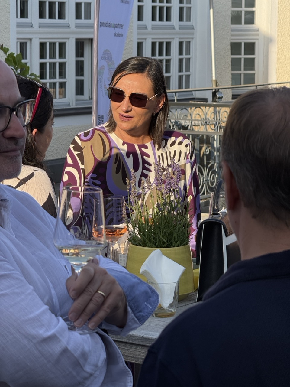 Das Bild steht in Zusammenhang mit Mediation und Konfliktlösung durch die Kanzlei Ponschab & Partner und zeigt folgendes: Eine Frau mit Sonnenbrille und gemustertem Hemd sitzt mit drei anderen Personen an einem Tisch im Freien, umgeben von Weingläsern und einer Lavendelpflanze im Topf. Das Sonnenlicht beleuchtet ihr Gesicht und ihren Oberkörper. Bei Fragen zu Konflikten und Konfliktlösung wenden Sie sich bitte an uns unter info@ponschab-partner.com