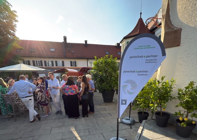 Das Bild steht in Zusammenhang mit Mediation und Konfliktlösung durch die Kanzlei Ponschab & Partner und zeigt folgendes: Eine Gruppe von Menschen steht und unterhält sich auf einer sonnigen Terrasse neben Topfpflanzen. Auf einem Schild im Vordergrund steht "ponschab & partner kanzlei für mediation" und "ponschab & partner akademie". Im Hintergrund sind Gebäude zu sehen. Bei Fragen zu Konflikten und Konfliktlösung wenden Sie sich bitte an uns unter info@ponschab-partner.com