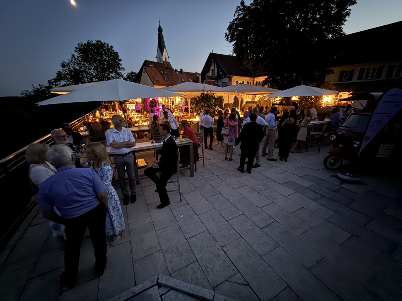Das Bild steht in Zusammenhang mit Mediation und Konfliktlösung durch die Kanzlei Ponschab & Partner und zeigt folgendes: Menschen versammeln sich bei einer Abendveranstaltung im Freien mit Tischen, Schirmen und warmer Beleuchtung. Einige stehen und unterhalten sich, während andere sitzen. Im Hintergrund sind Gebäude mit beleuchteten Fenstern und ein Kirchturm zu sehen. Bei Fragen zu Konflikten und Konfliktlösung wenden Sie sich bitte an uns unter info@ponschab-partner.com