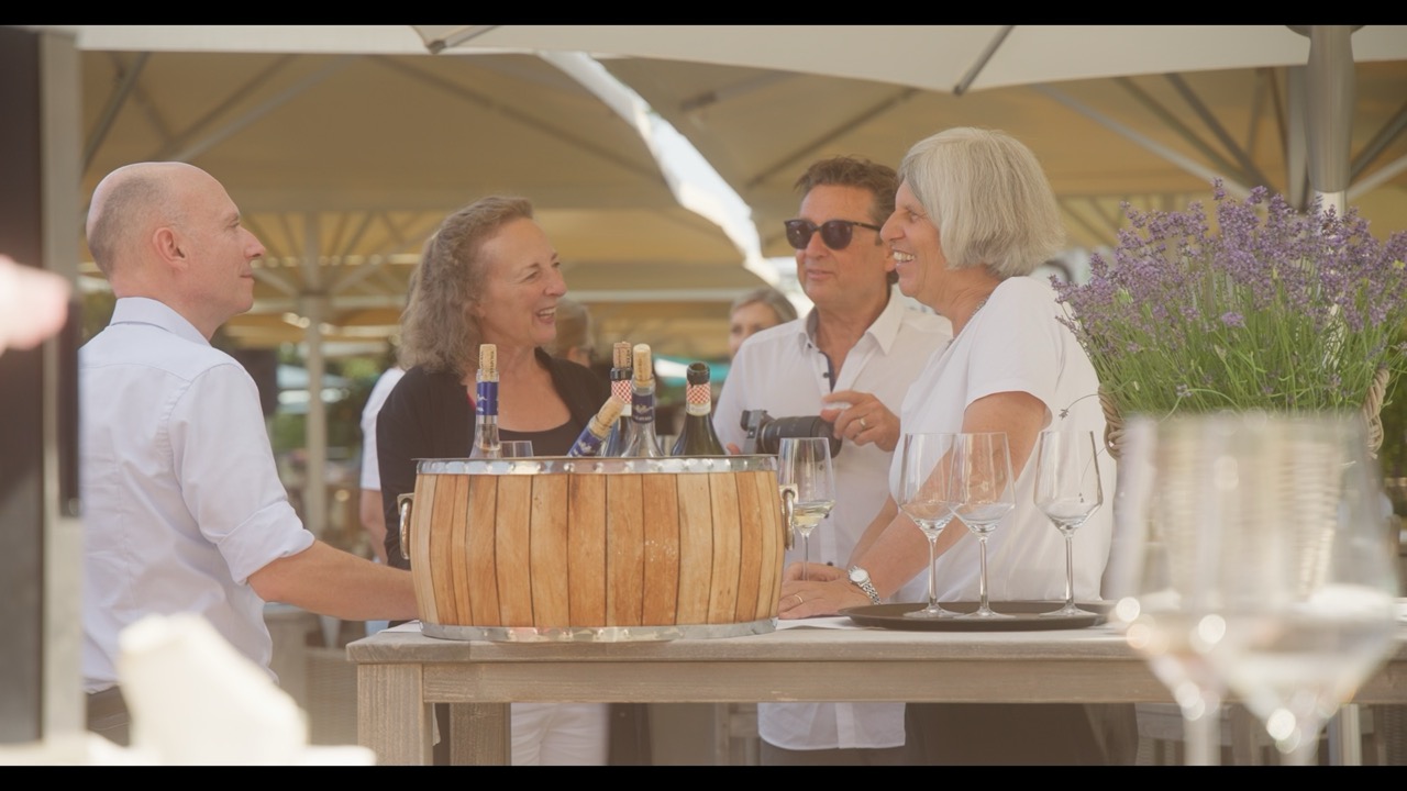 Das Bild steht in Zusammenhang mit Mediation und Konfliktlösung durch die Kanzlei Ponschab & Partner und zeigt folgendes: Vier Erwachsene stehen um einen Tisch im Freien, lächeln und unterhalten sich. Ein Eiskübel mit Weinflaschen steht zusammen mit leeren Weingläsern auf dem Tisch. Große Sonnenschirme spenden Schatten im Hintergrund. Bei Fragen zu Konflikten und Konfliktlösung wenden Sie sich bitte an uns unter info@ponschab-partner.com