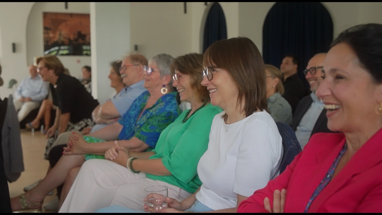 Das Bild steht in Zusammenhang mit Mediation und Konfliktlösung durch die Kanzlei Ponschab & Partner und zeigt folgendes: Eine Gruppe Erwachsener sitzt in einem Innenraum in Reihen, lächelt und lacht, während sie etwas beobachten, das sie nicht sehen können. Die meisten Menschen tragen Freizeit- oder Business-Casual-Kleidung und scheinen mit der Veranstaltung beschäftigt zu sein. Bei Fragen zu Konflikten und Konfliktlösung wenden Sie sich bitte an uns unter info@ponschab-partner.com