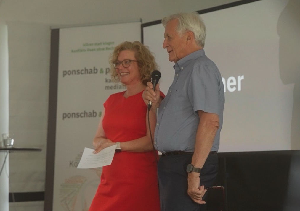 Das Bild steht in Zusammenhang mit Mediation und Konfliktlösung durch die Kanzlei Ponschab & Partner und zeigt folgendes: Eine Frau in einem roten Kleid hält Papiere in der Hand und steht neben einem älteren Mann in einem blauen Hemd, der ein Mikrofon hält. Sie sprechen vor einem Bildschirm und einem Banner mit der Aufschrift ponschab & partner. Bei Fragen zu Konflikten und Konfliktlösung wenden Sie sich bitte an uns unter info@ponschab-partner.com