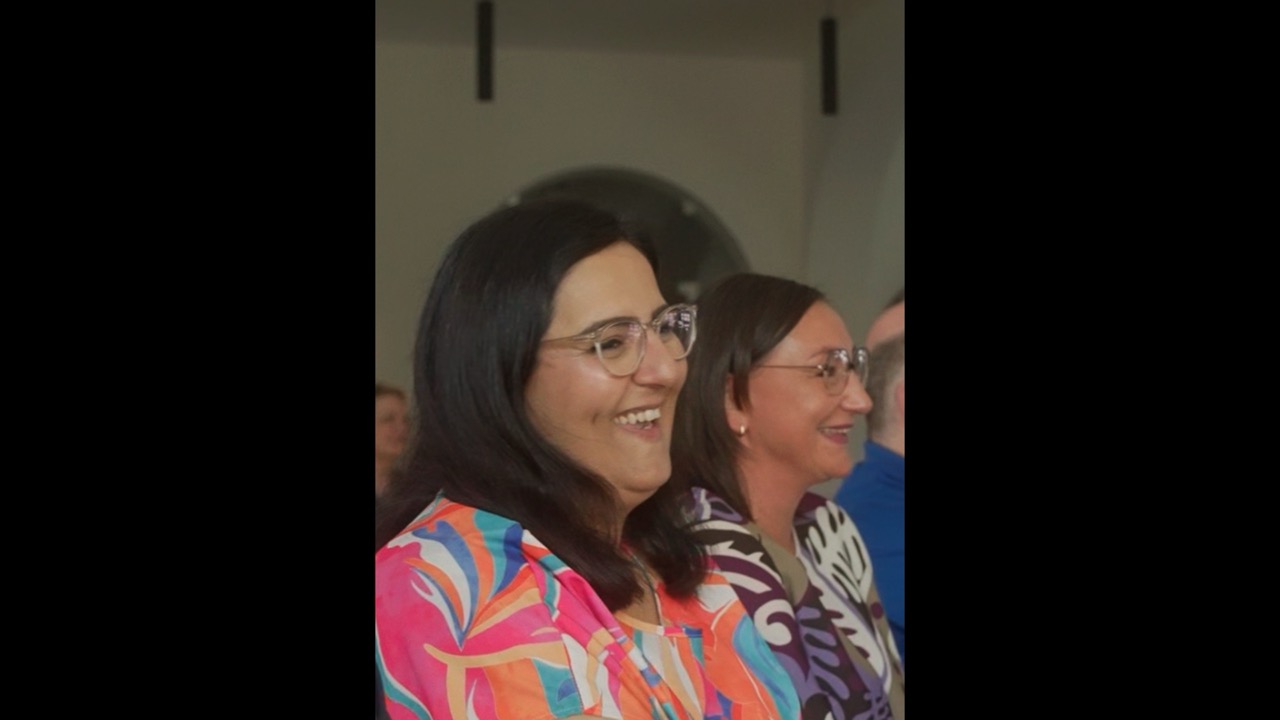 Das Bild steht in Zusammenhang mit Mediation und Konfliktlösung durch die Kanzlei Ponschab & Partner und zeigt folgendes: Zwei Frauen mit langen dunklen Haaren und Brillen sitzen nebeneinander, lächeln und schauen nach vorne. Beide tragen bunte, gemusterte Oberteile. Der Hintergrund ist unscharf und zeigt andere Menschen und ein rundes Fenster. Bei Fragen zu Konflikten und Konfliktlösung wenden Sie sich bitte an uns unter info@ponschab-partner.com