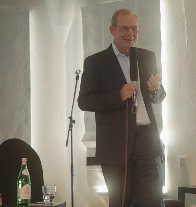 Das Bild steht in Zusammenhang mit Mediation und Konfliktlösung durch die Kanzlei Ponschab & Partner und zeigt folgendes: Ein Mann im Anzug steht in einem Raum und hält ein Mikrofon in der Hand, während er lächelnd spricht. Ein Mikrofonständer, ein Glas und eine Flasche San-Pellegrino-Wasser stehen auf einem kleinen Tisch in der Nähe. Von hinten dringt Licht ein. Bei Fragen zu Konflikten und Konfliktlösung wenden Sie sich bitte an uns unter info@ponschab-partner.com