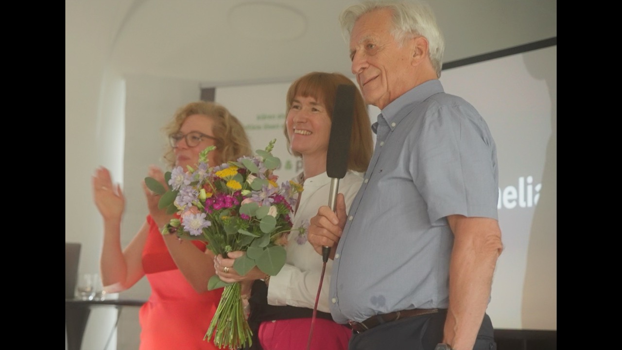 Das Bild steht in Zusammenhang mit Mediation und Konfliktlösung durch die Kanzlei Ponschab & Partner und zeigt folgendes: Drei Erwachsene stehen in einem Innenraum zusammen. Die Person auf der linken Seite klatscht, die Person in der Mitte hält einen Blumenstrauß und lächelt, und die Person auf der rechten Seite hält ein Mikrofon. Im Hintergrund ist ein Präsentationsbildschirm zu sehen. Bei Fragen zu Konflikten und Konfliktlösung wenden Sie sich bitte an uns unter info@ponschab-partner.com