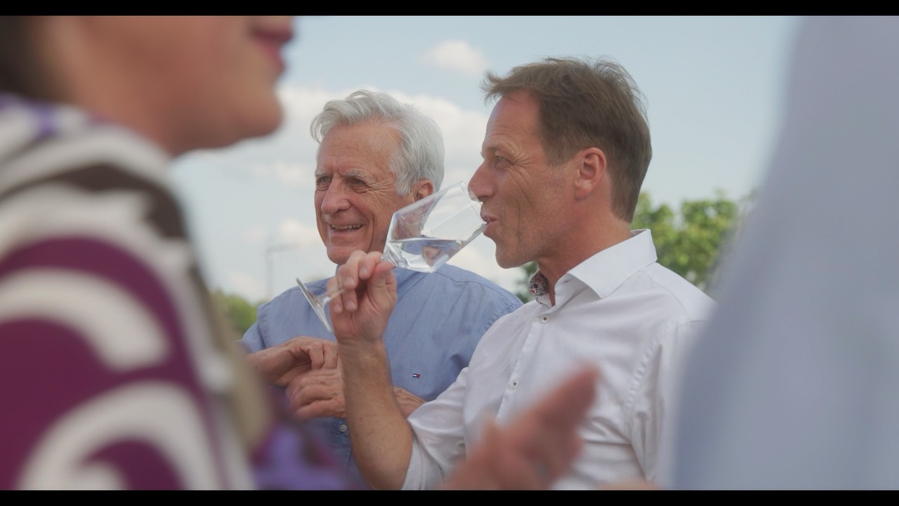 Das Bild steht in Zusammenhang mit Mediation und Konfliktlösung durch die Kanzlei Ponschab & Partner und zeigt folgendes: Zwei Männer in hellen Hemden stehen im Freien, halten Cocktailgläser in der Hand und trinken daraus. Der Vordergrund ist unscharf und zeigt einen Teil einer Person in gemusterter Kleidung. Der Hintergrund ist ein sonniger Himmel mit etwas Grün. Bei Fragen zu Konflikten und Konfliktlösung wenden Sie sich bitte an uns unter info@ponschab-partner.com