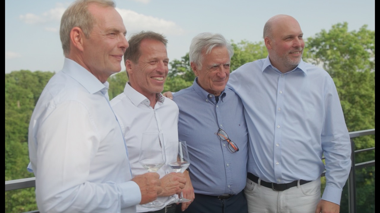 Das Bild steht in Zusammenhang mit Mediation und Konfliktlösung durch die Kanzlei Ponschab & Partner und zeigt folgendes: Vier Männer in hellen Hemden stehen zusammen auf einem Balkon und lächeln in die Kamera. Zwei halten Weingläser in der Hand. Im Hintergrund sind Bäume und ein blauer Himmel mit Wolken zu sehen. Bei Fragen zu Konflikten und Konfliktlösung wenden Sie sich bitte an uns unter info@ponschab-partner.com