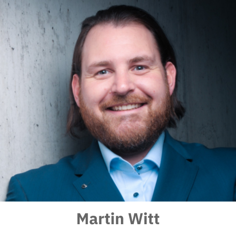 Das Bild steht in Zusammenhang mit Mediation und Konfliktlösung durch die Kanzlei Ponschab & Partner und zeigt folgendes: Ein Mann mit langen braunen Haaren und einem Bart, der eine blaue Anzugsjacke und ein hellblaues Hemd trägt, lächelt vor einer Betonwand in die Kamera. Der Text Martin Witt steht unter dem Bild. Bei Fragen zu Konflikten und Konfliktlösung wenden Sie sich bitte an uns unter info@ponschab-partner.com