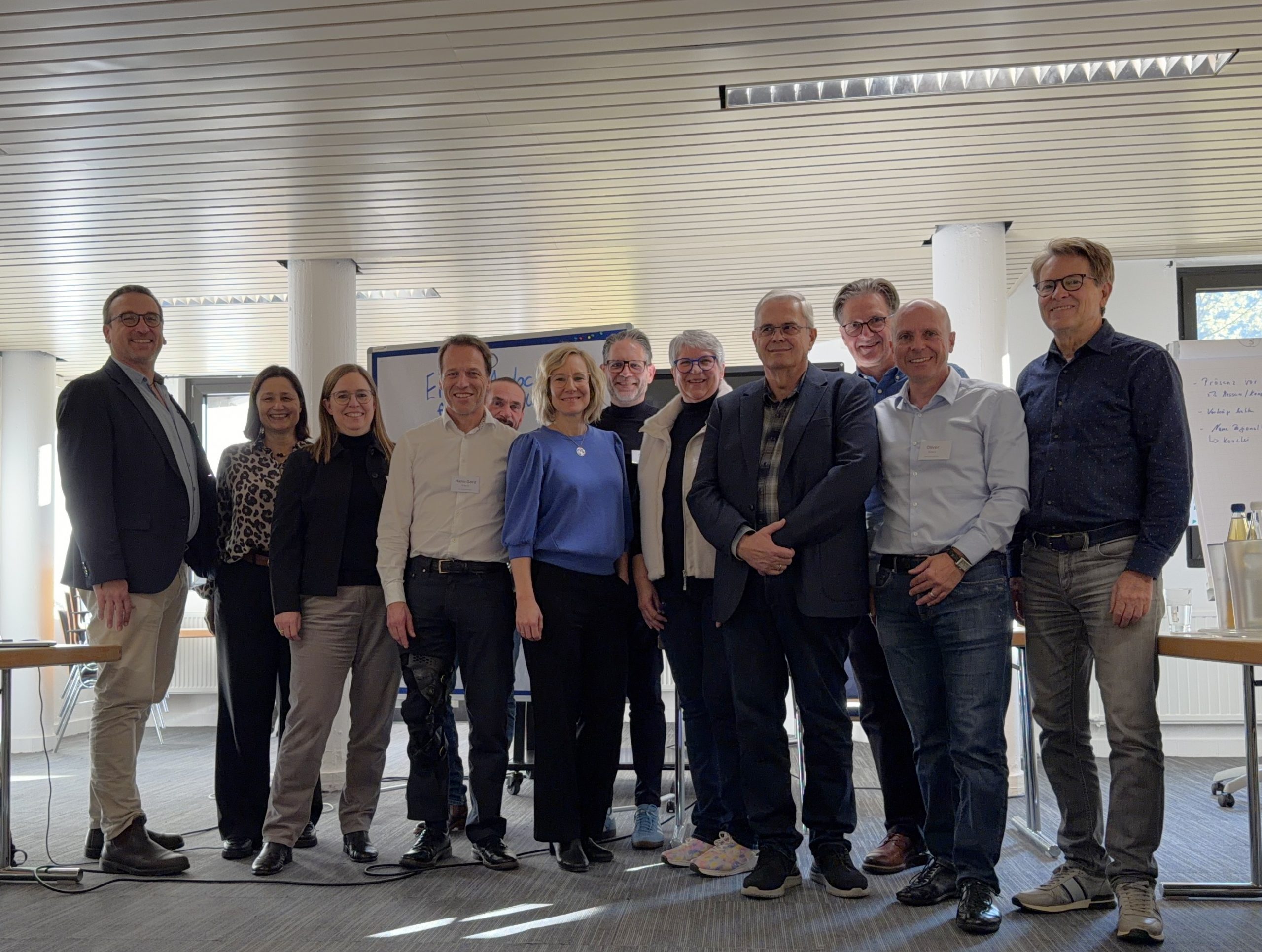 Das Bild steht in Zusammenhang mit Mediation und Konfliktlösung durch die Kanzlei Ponschab & Partner und zeigt folgendes: Eine Gruppe von elf Erwachsenen steht drinnen in einem gut beleuchteten Konferenzraum zusammen. Sie sind in legerer Business-Kleidung gekleidet und lächeln in die Kamera. Im Hintergrund sind Tische, Stühle und ein Flipchart zu sehen. Bei Fragen zu Konflikten und Konfliktlösung wenden Sie sich bitte an uns unter info@ponschab-partner.com